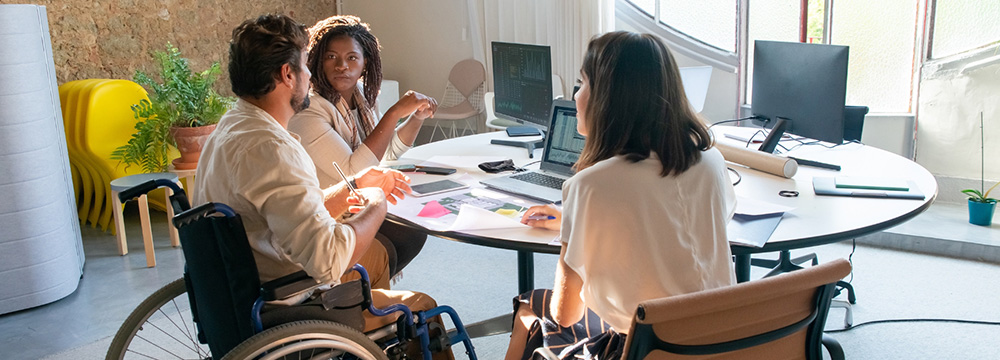 Three people around a laptop. One person is in a wheelchair. 